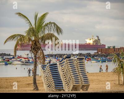 Am Strand stapelten sich Liegestühle, im Hintergrund ein Containerschiff und Boote im Wasser, Sommeratmosphäre, teneriffa, kanarische Inseln, spanien Stockfoto