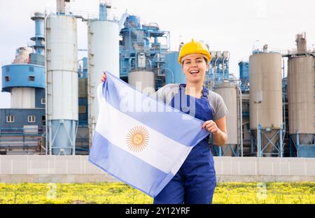 Junge Ingenieurin im Helm, die die Staatsflagge Argentiniens winkte, während sie vor großen Tanks in der Chemiefabrik stand Stockfoto