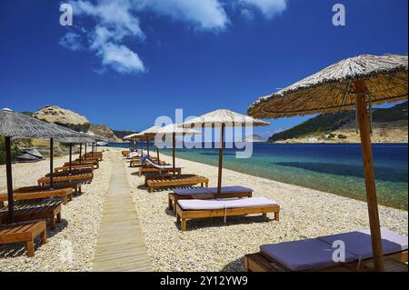 Liegestühle und Sonnenschirme am Strand mit Holzsteg, freier Blick auf das ruhige Meer und den blauen Himmel, Petras Beach, Kalikatsou Rock, Patmos, Dodekanese, Stockfoto