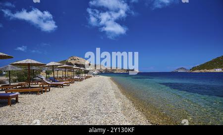 Ein leerer Strand mit Liegestühlen und Strohschirmen auf dem klaren blauen Wasser eines Meeres unter einem hellblauen Himmel, Petras Beach, Kalikatsou Rock, Patmos, D Stockfoto