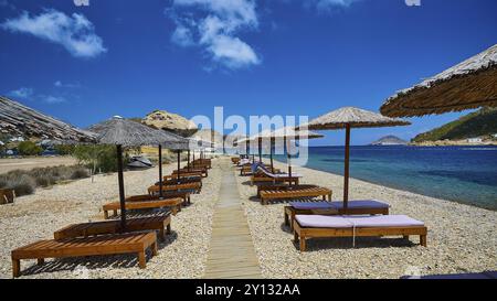 Ein Strand mit einem Holzsteg und Liegestühlen unter Strohschirmen, der Blick auf das klare Wasser und den sonnigen Himmel, Petras Beach, Kalikatsou Rock, P Stockfoto