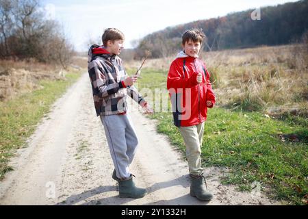 Zwei Jungs laufen auf einem unbefestigten Weg, jeder hält einen Stock. Sie sind lässig für einen sonnigen Tag gekleidet, umgeben von der Schönheit der Natur im frühen Herbst. Stockfoto