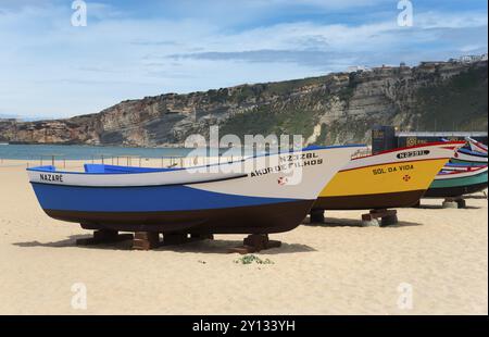 Mehrere farbenfrohe Fischerboote liegen am Sandstrand von Nazare mit Hügeln im Hintergrund, Praia da Nazare Strand, Nazare, Oeste, Bezirk Leiria Stockfoto