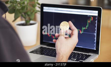 Mann, der Kryptowährungsdiagramme und Bitcoin-Münzen auf einem Laptop überprüft Stockfoto