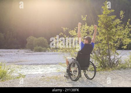 Glücklicher und fröhlicher junger Mann im Rollstuhl draußen in der Natur an einem sonnigen Sommertag mit erhobenen Händen Stockfoto