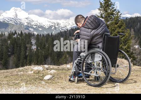 Foto eines Mannes im Rollstuhl mit Kamera in der Natur, der wunderschöne Berge fotografiert Stockfoto