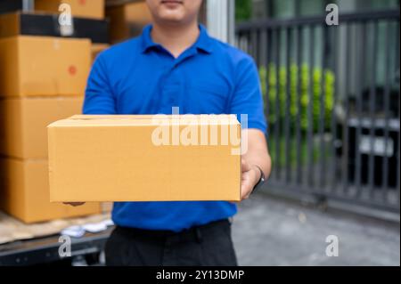 Ein zugeschnittenes Bild eines asiatischen Zustellers in blauer Uniform, der hinten am Lkw steht und ein Paket zur Lieferung an einen Kunden trägt. Versand Stockfoto