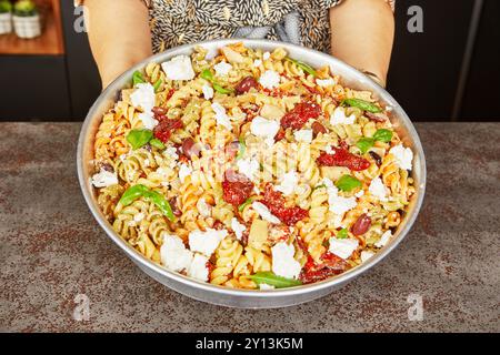 Eine große Schüssel mediterraner Nudelsalat mit dreifarbigen Fusilli, Fetakäse, sonnengetrockneten Tomaten, Oliven und frischen Basilikumblättern, gehalten von einem Pers Stockfoto