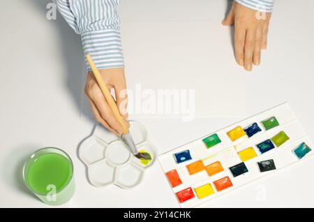 Künstler mischt Aquarellfarben in Keramikpalette im Atelier, Nahaufnahme. Weibliche Hände und Aquarellzubehör auf dem Tisch, Draufsicht Stockfoto