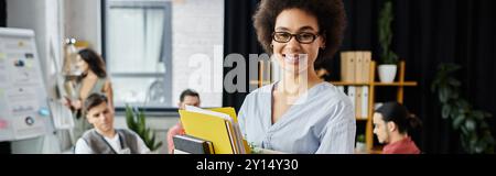 Fröhliche afroamerikanische Frau, die ihre Sachen während der Entlassung packt, Kollegen im Hintergrund. Stockfoto