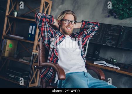 Foto eines fröhlichen bärtigen Programmierers, junger Mann, der im Büro auf einem Sessel sitzt und auf einem Computerbildschirm sitzt Stockfoto