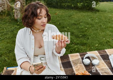 Eine junge Frau in einem weißen Hemd genießt Croissant, während sie sich an einem sonnigen Sommertag im Freien entspannt. Stockfoto