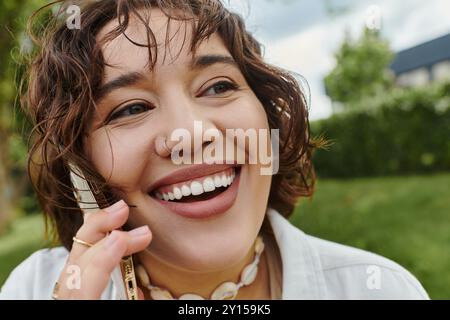 Eine fröhliche junge Frau in einem weißen Hemd entspannt sich in der Sommersonne und plaudert fröhlich auf ihrem Telefon Stockfoto