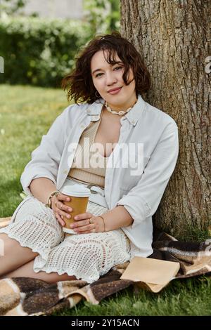 Eine junge Frau genießt einen sonnigen Tag in einem grünen Park, schlürft Kaffee und entspannt sich unter einem Baum. Stockfoto