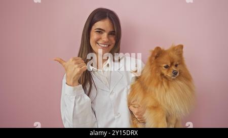 Eine lächelnde junge Frau zeigt auf ihre Seite, während sie einen pommerschen Hund an eine rosa Wand hält. Stockfoto