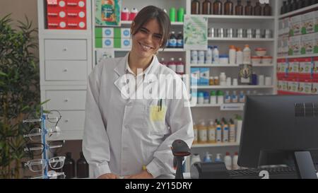 Eine lächelnde junge Frau in einer Apotheke, die einen weißen Labormantel trägt, steht neben Brillen und Gesundheitsprodukten. Stockfoto