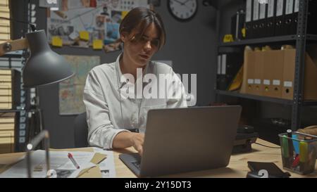 Fokussierte junge Frau, die an einem Laptop arbeitet, in einem schwach beleuchteten Detektivbüro, umgeben von Beweisen. Stockfoto