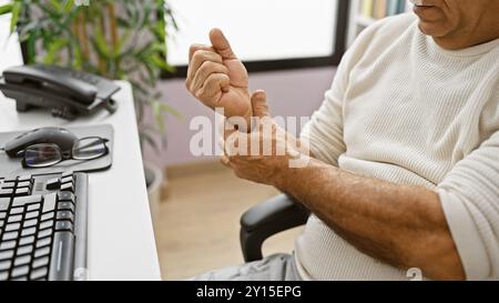 Reifer hispanischer Mann im Büro, der Handgelenkschmerzen spürt, mit Computer, Brille, Telefon und Pflanze im Hintergrund Stockfoto