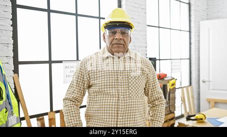 Ein reifer hispanischer Mann in Sicherheitsausrüstung steht selbstbewusst in einer gut ausgestatteten Tischlerei. Stockfoto