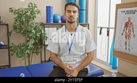 Ein gutaussehender hispanischer Mann, der einen weißen Mantel und ein Schlüsselband trägt, posiert selbstbewusst in einer hellen Rehabilitationsklinik. Stockfoto