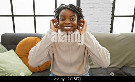 Eine lächelnde junge schwarze Frau mit Dreadlocks, die ihre Ohren bedecken, während sie drinnen auf einem Sofa sitzt. Stockfoto
