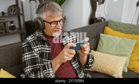 Ein fröhlicher Mann mittleren Alters mit Kopfhörern benutzt ein Smartphone in einem gemütlichen Wohnzimmer, was auf moderne Freizeit hinweist. Stockfoto
