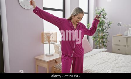 Die junge kaukasische Frau tanzt fröhlich im Pyjama mit Smartphone in ihrem gemütlichen Schlafzimmer. Stockfoto