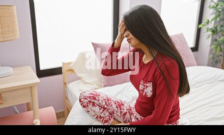 Gestresste indische Frau mit langen Haaren, die in einem Schlafzimmer sitzt, Pyjamas trägt und den Kopf hält. Stockfoto
