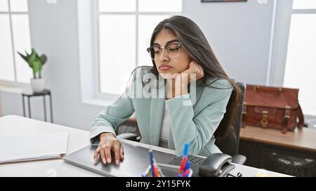 Gelangweilte indianerin in Brille, die an ihrem Schreibtisch sitzt und ein gemeinsames Szenario am Arbeitsplatz darstellt. Stockfoto