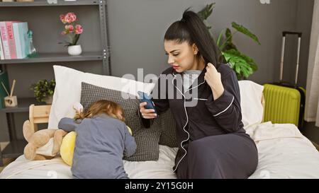 Eine junge Frau überprüft ihr Handy, während ihre Tochter neben ihr auf einem Bett in einem modern eingerichteten Schlafzimmer schläft. Stockfoto