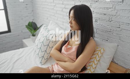 Eine junge chinesin sitzt auf dem Bett in einem modernen Schlafzimmer mit einer Pflanze im Hintergrund, sieht ernst aus und trägt ein pinkfarbenes Satinoberteil Stockfoto