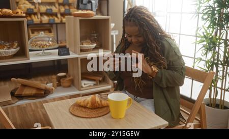 Frau fotografiert ein Croissant und Kaffee auf einem Tisch in einer gemütlichen Bäckerei mit Brotregalen im Hintergrund Stockfoto