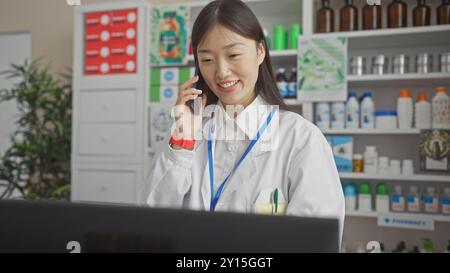 Eine lächelnde junge asiatische Apothekerin in einem weißen Mantel unterhält sich auf einem Smartphone in einer modernen Apotheke. Stockfoto