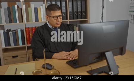 Junger hispanischer Mann in einem Richtergewand, der an einem Computer arbeitet, in einem gut organisierten Büro mit Bücherregalen Stockfoto