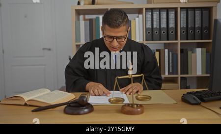 Junger hispanischer Mann in Richterbekleidung liest Dokumente in einem Büro mit Rechtsbüchern und Justizskalen auf dem Schreibtisch Stockfoto