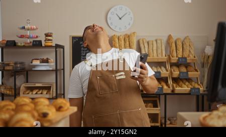 Junger hispanischer Mann in einer Bäckerei mit Schürze, lächelnd, während er ein Smartphone hält, mit verschiedenen Broten und Gebäck im Hintergrund, aufgenommen in A Stockfoto