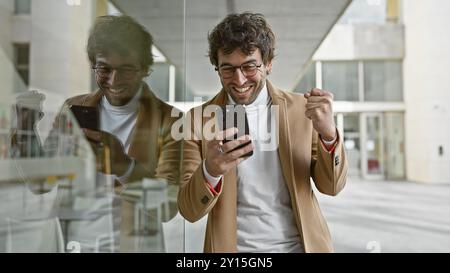 Ein fröhlicher hispanischer Mann feiert eine Nachricht auf seinem Smartphone auf einer Stadtstraße, die moderne urbane Konnektivität widerspiegelt. Stockfoto