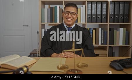 Junger hispanischer Mann, der an einem Schreibtisch in einem Büro sitzt, einen Richtermantel trägt, umgeben von Büchern und juristischen Dokumenten, und in einem Beruf selbstbewusst lächelt Stockfoto