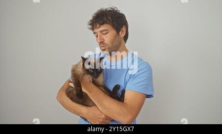 Junger hispanischer Mann, der eine siamesische Katze vor weißem Hintergrund hält Stockfoto