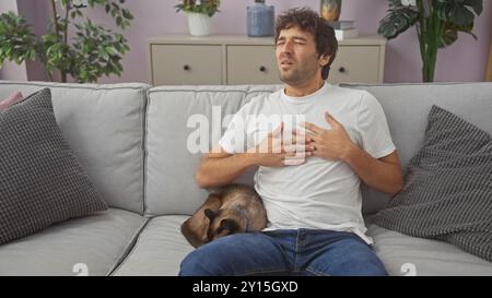 Ein junger Mann, der sich auf einer Couch unwohl fühlt, mit einer siamkatze, die neben ihm in einem gemütlichen Wohnzimmer ruht. Stockfoto