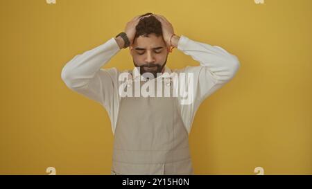 Ein verzweifelter hispanischer Mann in Schürze und Pullover steht mit geschlossenen Augen, Hände auf dem Kopf, an einer gelben Wand. Stockfoto