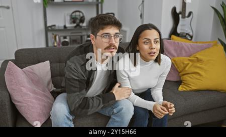 Ein Paar sitzt dicht auf einer Couch in einem modernen Wohnzimmer und sieht überrascht und aufmerksam aus. Stockfoto