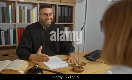 Männlicher Richter im Amt, der mit einem Klienten spricht, während Bücher, Waagen und Hammer mit Papieren auf dem Schreibtisch liegen Stockfoto