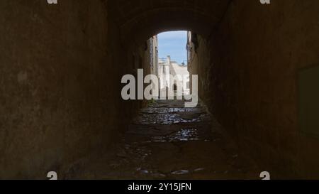 Eine enge, kopfsteingepflasterte Gasse in matera, basilicata, italien, bietet einen hellen Blick auf historische Steinhäuser unter einem klaren blauen Himmel durch einen Bogen Stockfoto