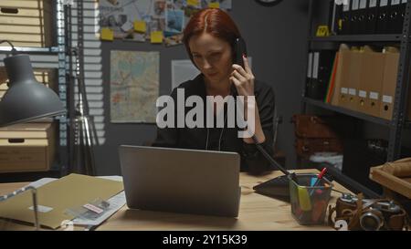 Ernsthafte rothaarige Detektivin im Büro mit Laptop und Telefon, umgeben von Beweismitteln und Ermittlungsausrüstung. Stockfoto