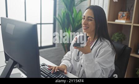 Hispanische Ärztin lacht, während sie eine Sprachnachricht an ihrem Krankenhausarbeitsplatz aufnimmt Stockfoto