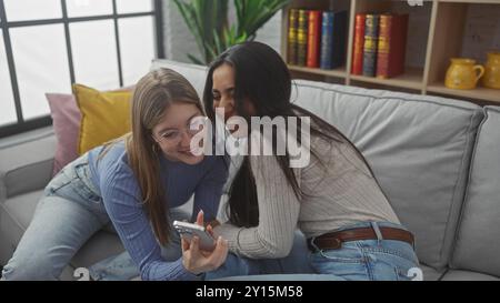 Zwei Frauen, die lachen und zusammen ein Smartphone auf einer Couch im Haus anschauen, mit Büchern im Hintergrund Stockfoto