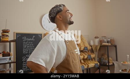 Junger, gutaussehender arabischer Mann mit einem Bart, der selbstbewusst lächelt, während er in einer Bäckerei mit Brot und Gebäck arbeitet. Stockfoto