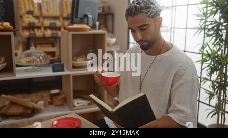 Junger arabischer Mann mit Bart liest ein Buch und trinkt Kaffee in einer Bäckerei mit Brot im Hintergrund. Stockfoto