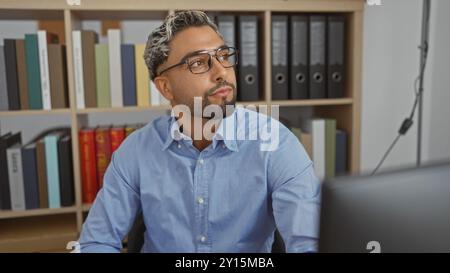 Junger, arabischer, muslimischer Mann mit Bart, der drinnen in einem Büroarbeitsplatz sitzt, umgeben von Regalen mit Büchern und Ordnern Stockfoto